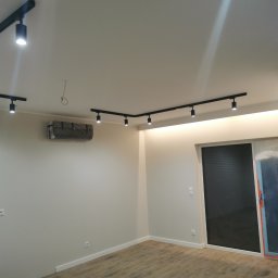 F.H.U.LUK- POL Łukasz Dereń - Pusty pokój z drewnianą podłogą, czarnym oświetleniem szynowym na suficie, klimatyzacją zabezpieczoną folią, oraz drzwiami przesuwnymi częściowo zakrytymi folią malarską.