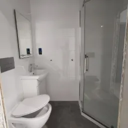 Nowoczesna, jasna łazienka z wiszącą toaletą z otwartą deską, umywalką na szafce, kabiną prysznicową z deszczownicą i szarą podłogą z płytek.