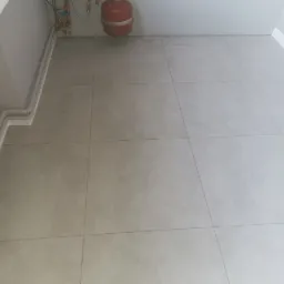 Ułożona jasnoszara podłoga z płytek ceramicznych z widocznymi fugami, grzejnik oraz rury CO przy ścianie, w tle listwa przypodłogowa.