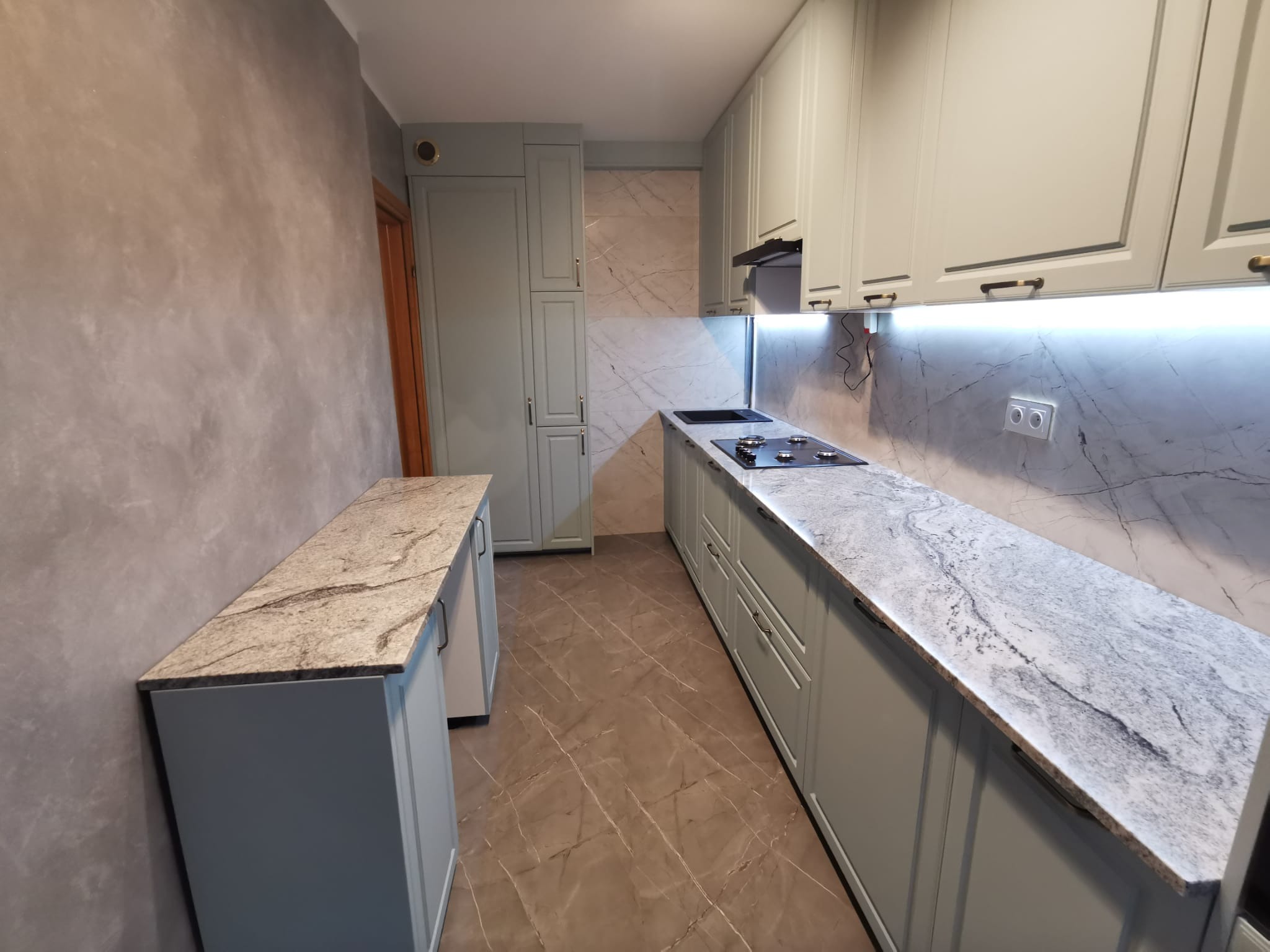 Nowoczesna kuchnia w odcieniach szarości z marmurowymi blatami i podświetleniem LED pod szafkami wiszącymi, widok na zabudowę kuchenną i wyspę.