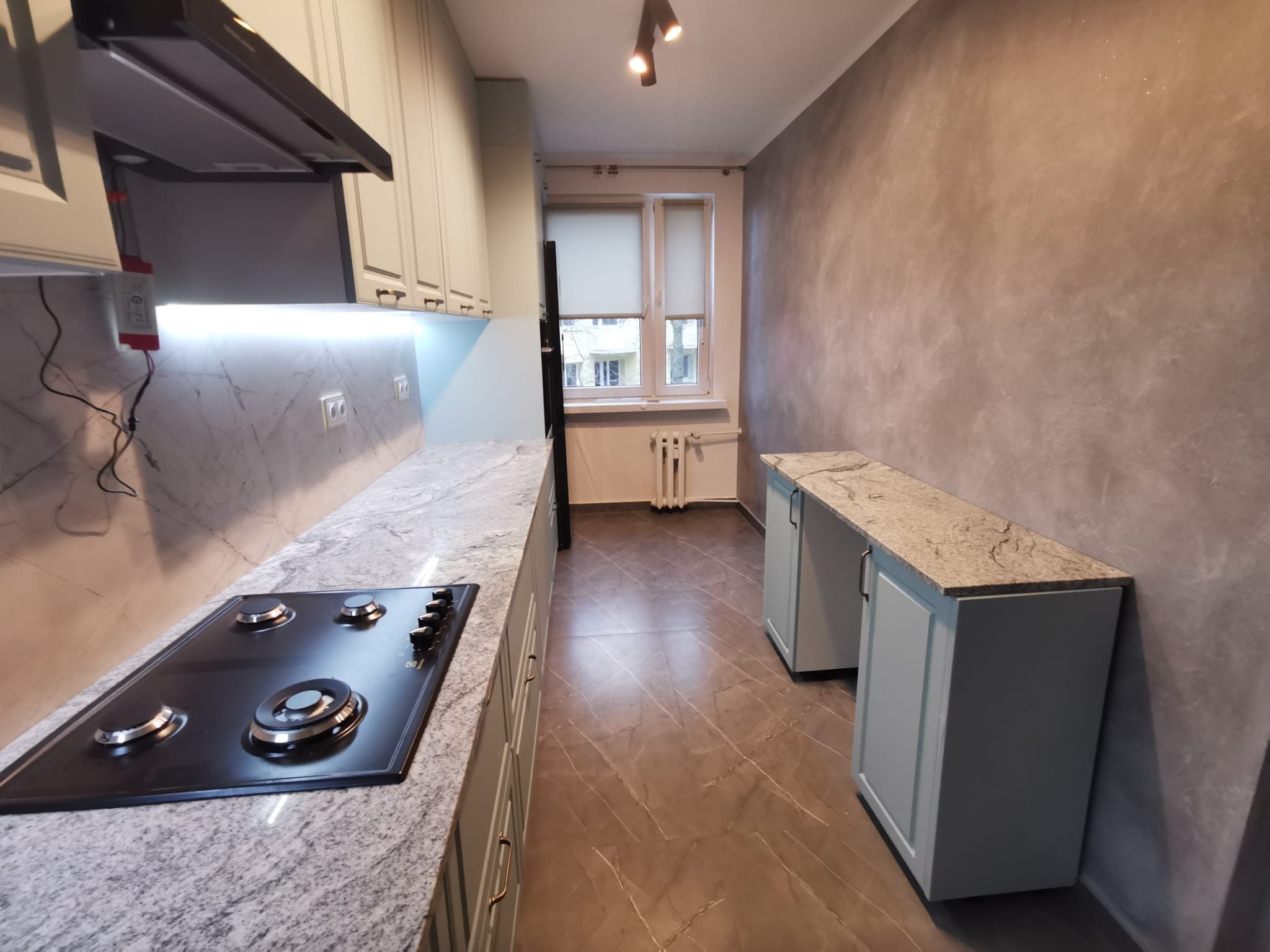 Nowoczesna kuchnia po remoncie: jasnoszare szafki z granitowym blatem, czarna płyta gazowa, ściana w odcieniach szarości z delikatnym połyskiem, brązowa podłoga imitująca marmur, okno z roletami.