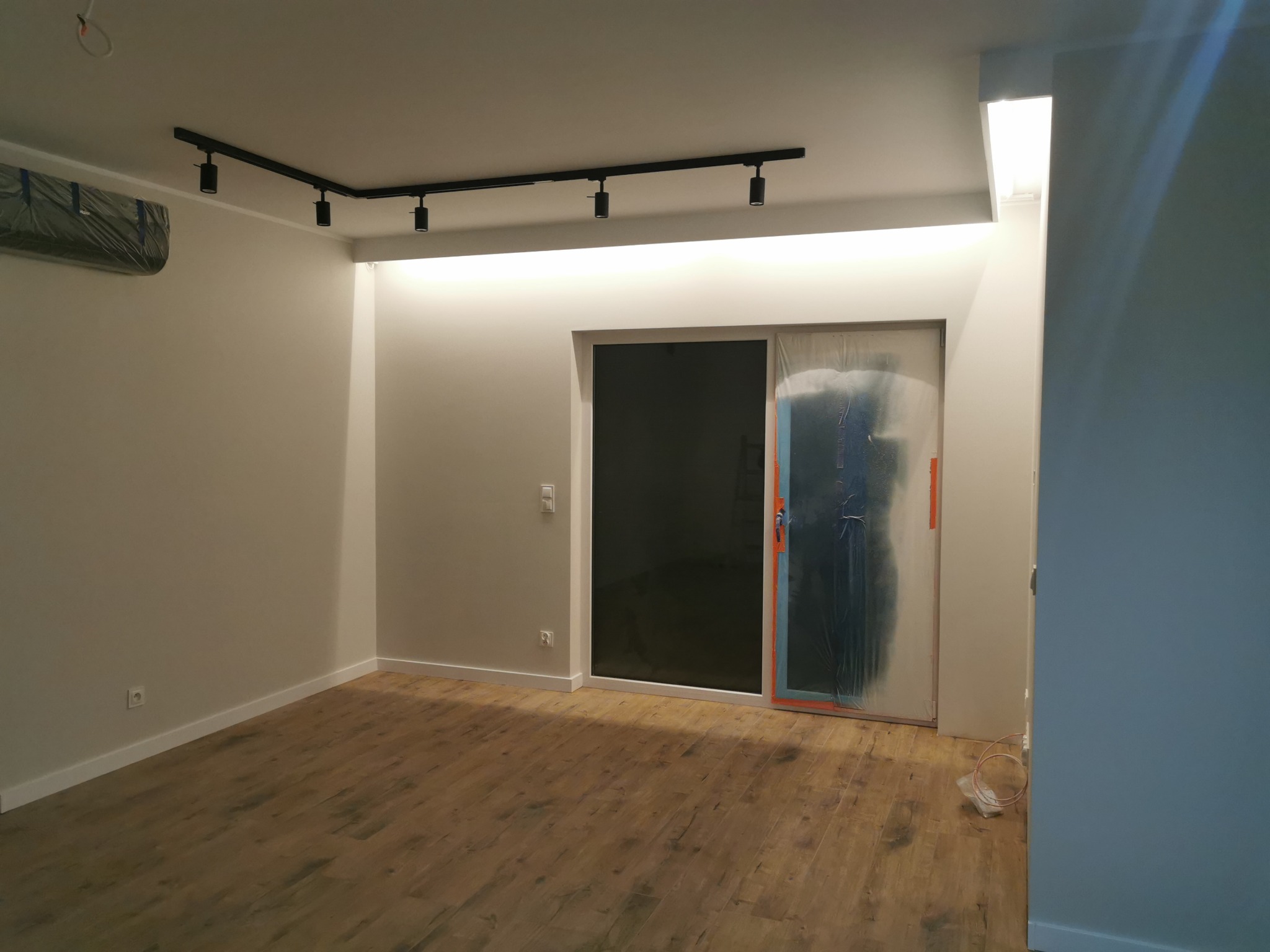 Wnętrze pokoju po remoncie z drewnianą podłogą, białymi ścianami, czarnym oświetleniem szynowym i osłoniętymi folią drzwiami przesuwnymi.