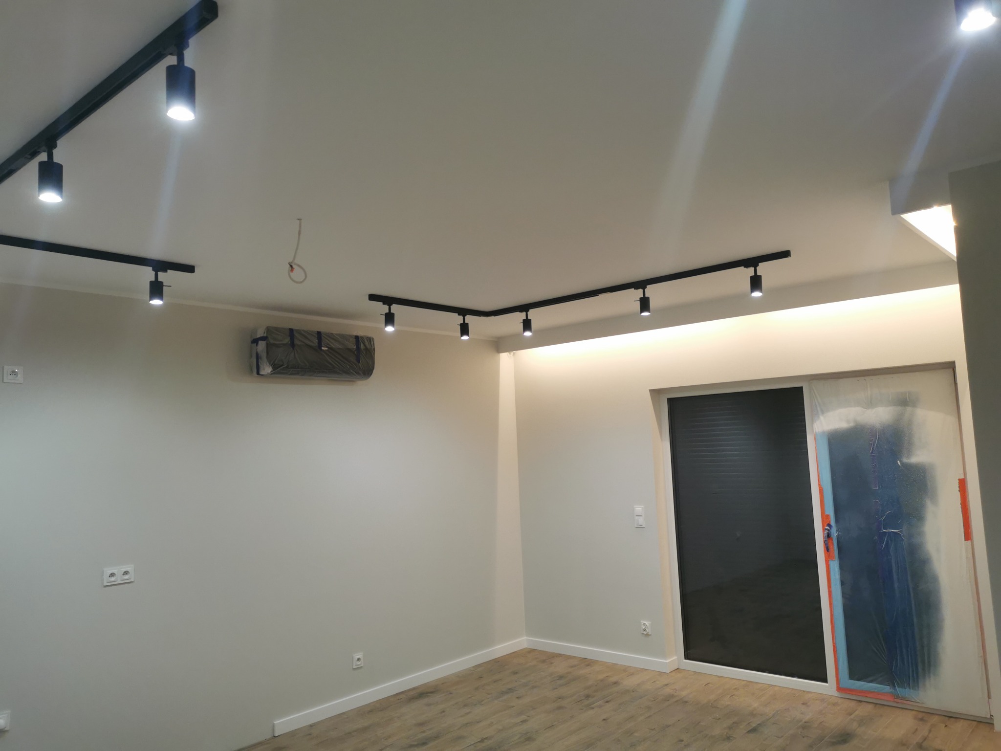 Pusty pokój z drewnianą podłogą, czarnym oświetleniem szynowym na suficie, klimatyzacją zabezpieczoną folią, oraz drzwiami przesuwnymi częściowo zakrytymi folią malarską.