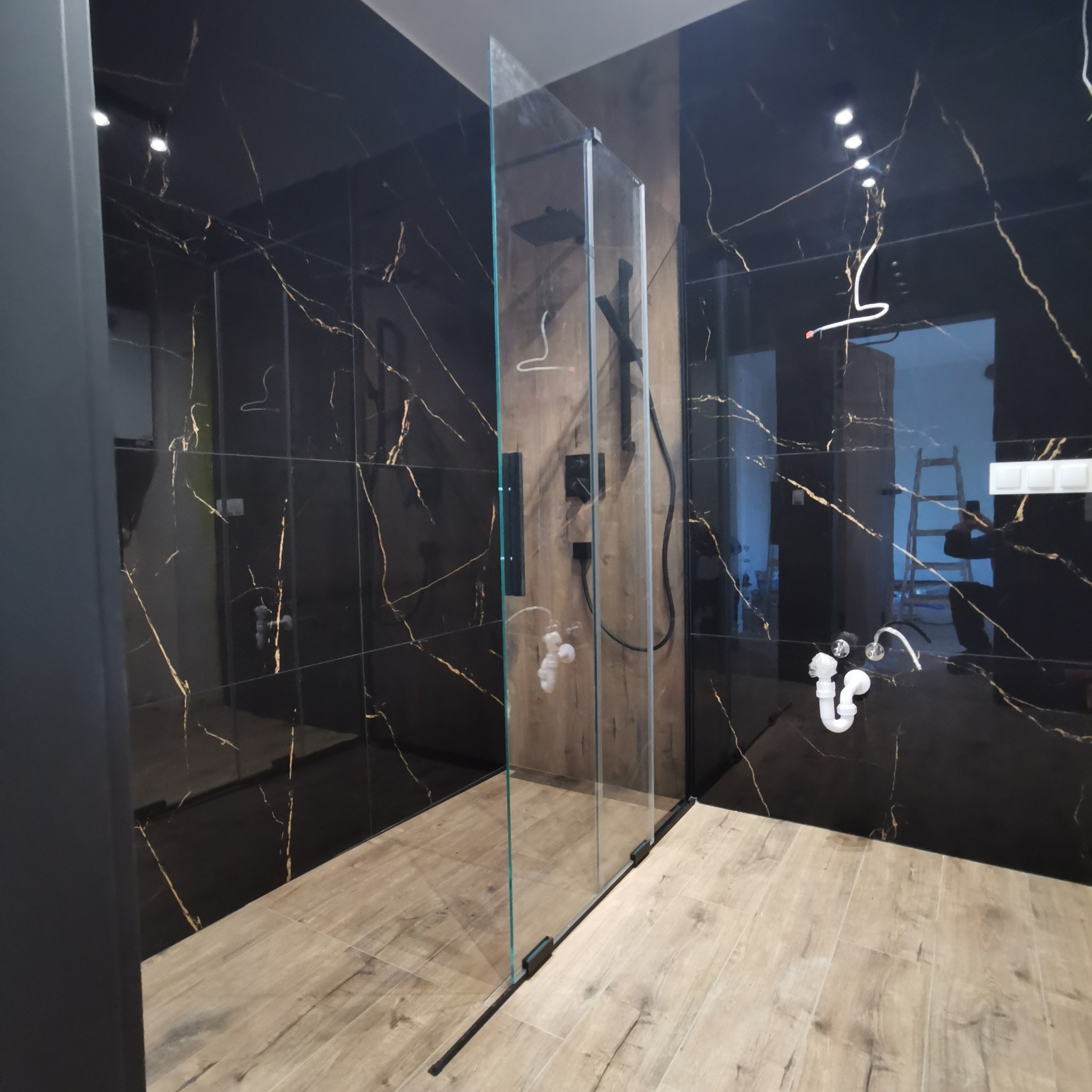 Nowoczesna łazienka z czarnymi płytkami imitującymi marmur, kabiną prysznicową typu walk-in z czarną armaturą i drewnopodobną podłogą, widoczne niedokończone podłączenia hydrauliczne.