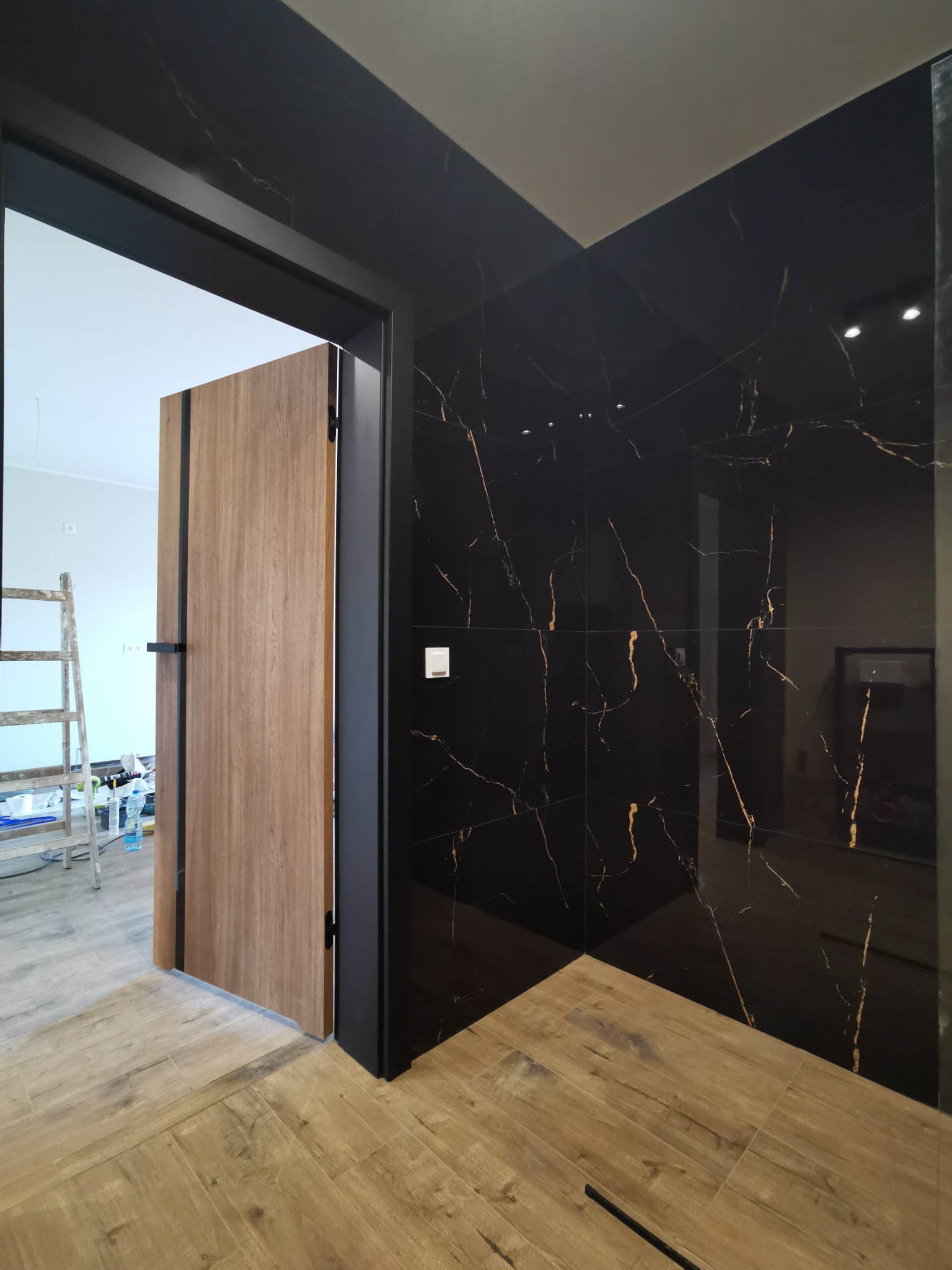 Częściowo otwarta, drewnopodobna, wewnętrzna stolarka drzwiowa z czarną ramą na tle ściany wyłożonej czarnymi płytkami z marmurowym wzorem, widoczny fragment podłogi wyłożonej płytkami...