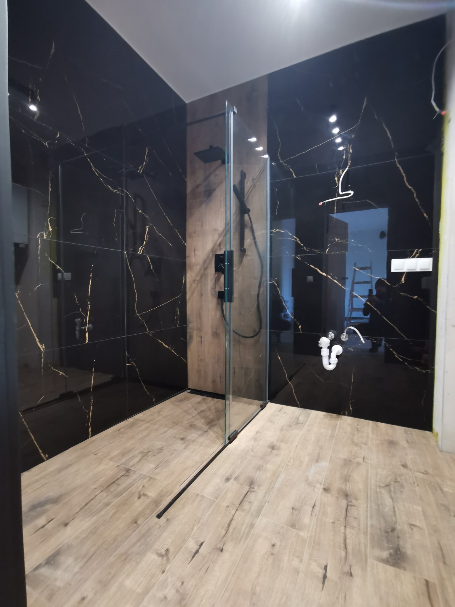 Nowoczesna łazienka z czarnymi płytkami imitującymi marmur ze złotymi żyłami, drewnopodobnymi płytkami podłogowymi i kabiną prysznicową typu walk-in z czarną armaturą, widoczne niedokończone prace...