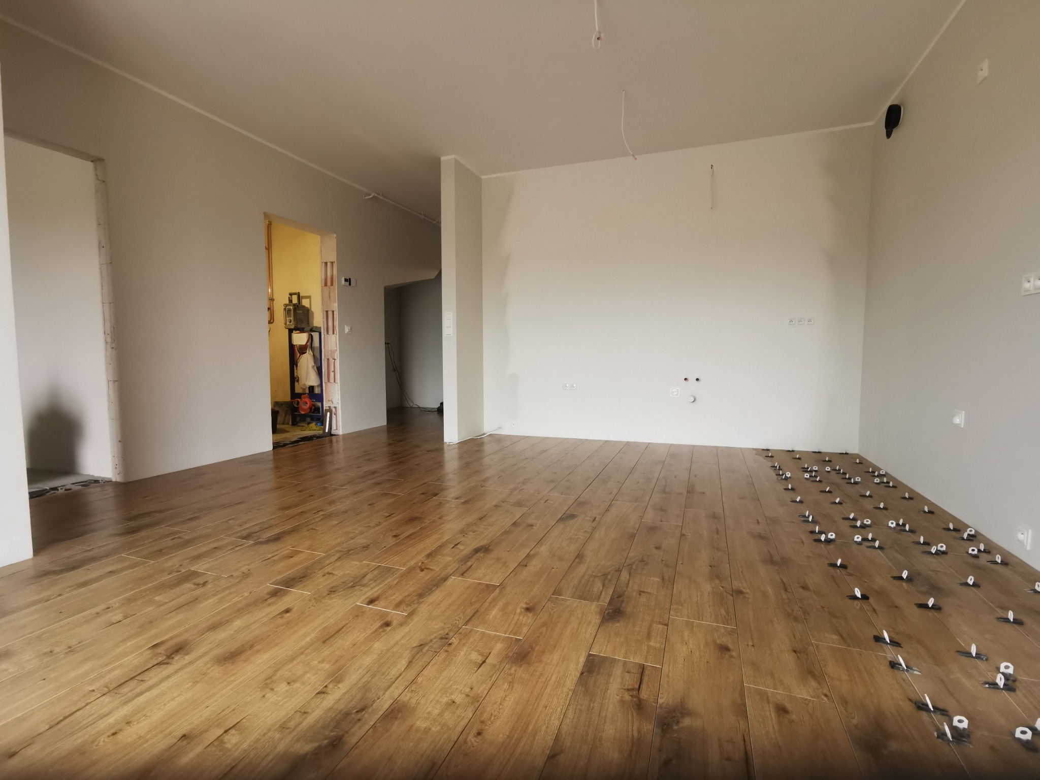 Układanie drewnopodobnych płytek podłogowych w przestronnym, jasnym pomieszczeniu z użyciem systemów poziomowania płytek; widoczne otwory na rury grzewcze w ścianie.