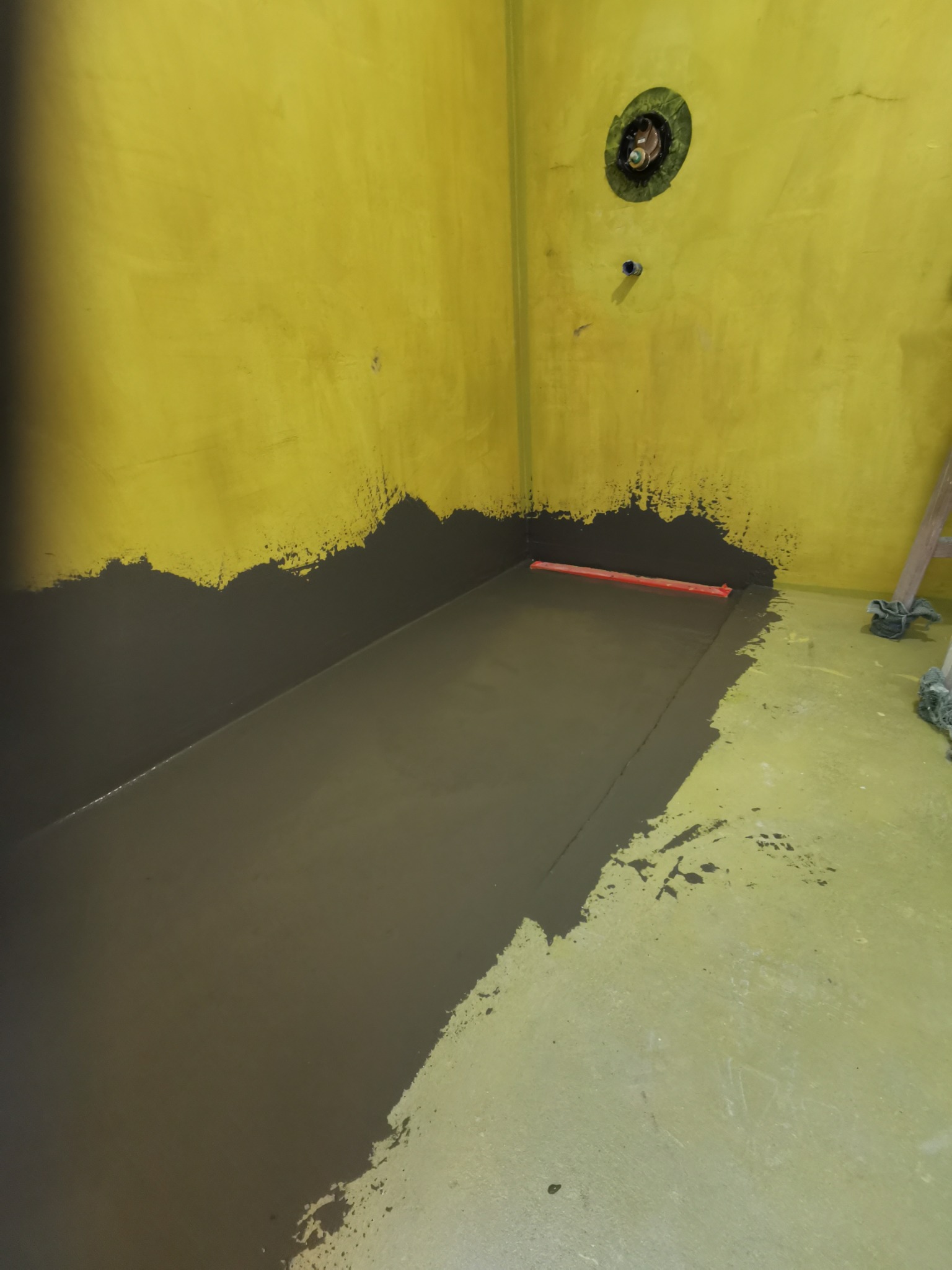 Przygotowanie podłoża w łazience: wylewka samopoziomująca na żółtej ścianie, widoczna poziomica i fragment drabiny.