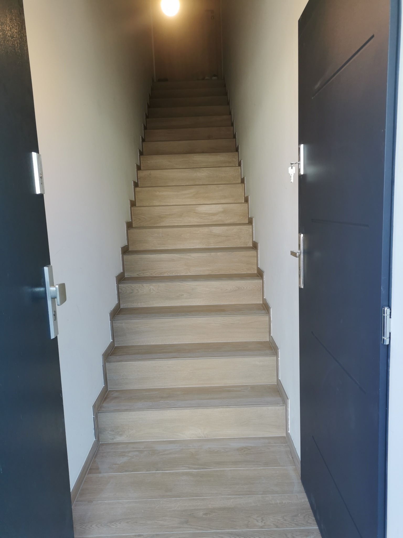 Jasne drewniane schody z widocznymi słojami prowadzące w górę, z fragmentami otwartych, ciemnych drzwi po bokach, oświetlone górnym światłem.