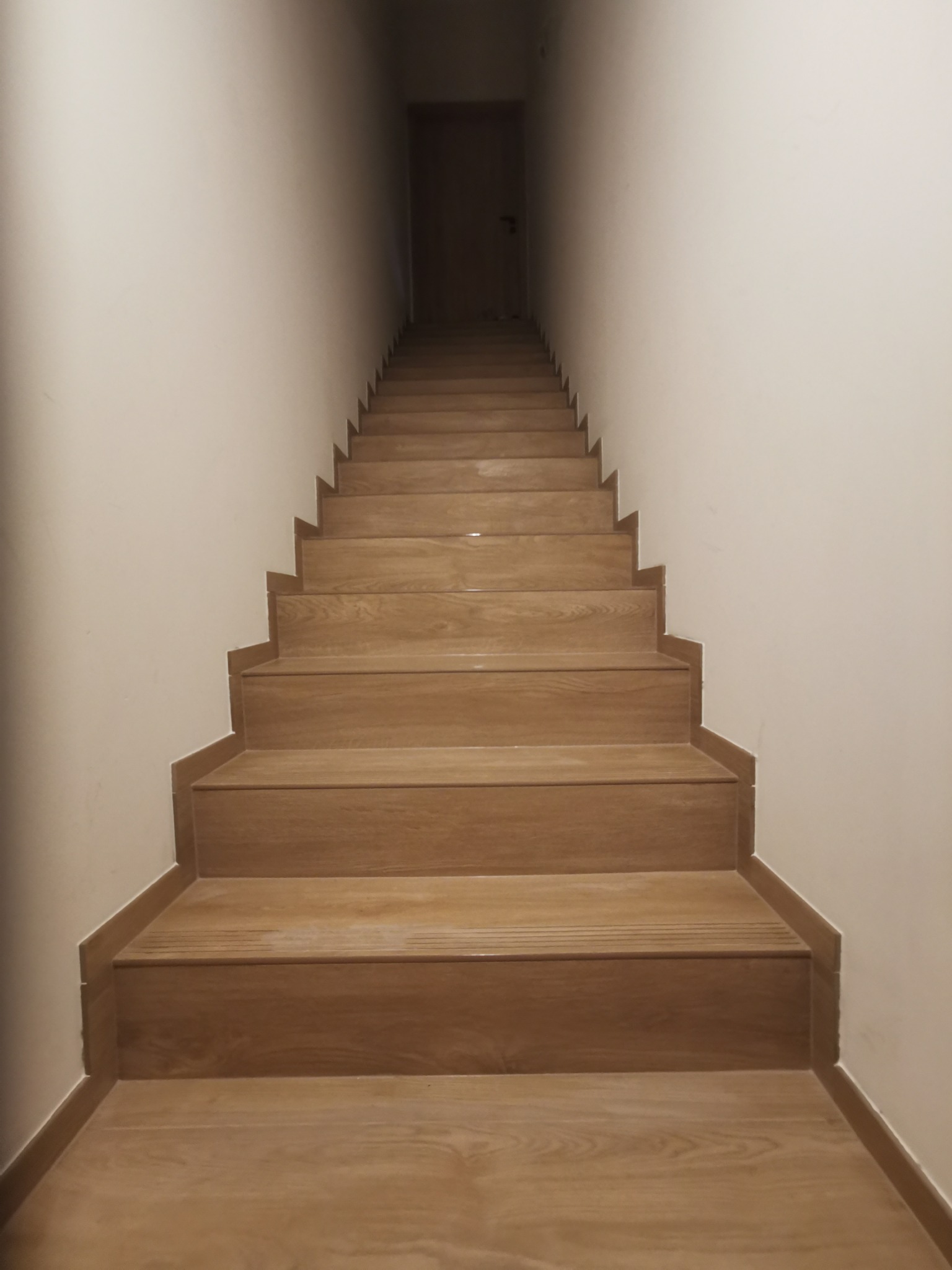 Schody wyłożone panelami imitującymi drewno, prowadzące do ciemnego pomieszczenia z drzwiami, widok z dołu.