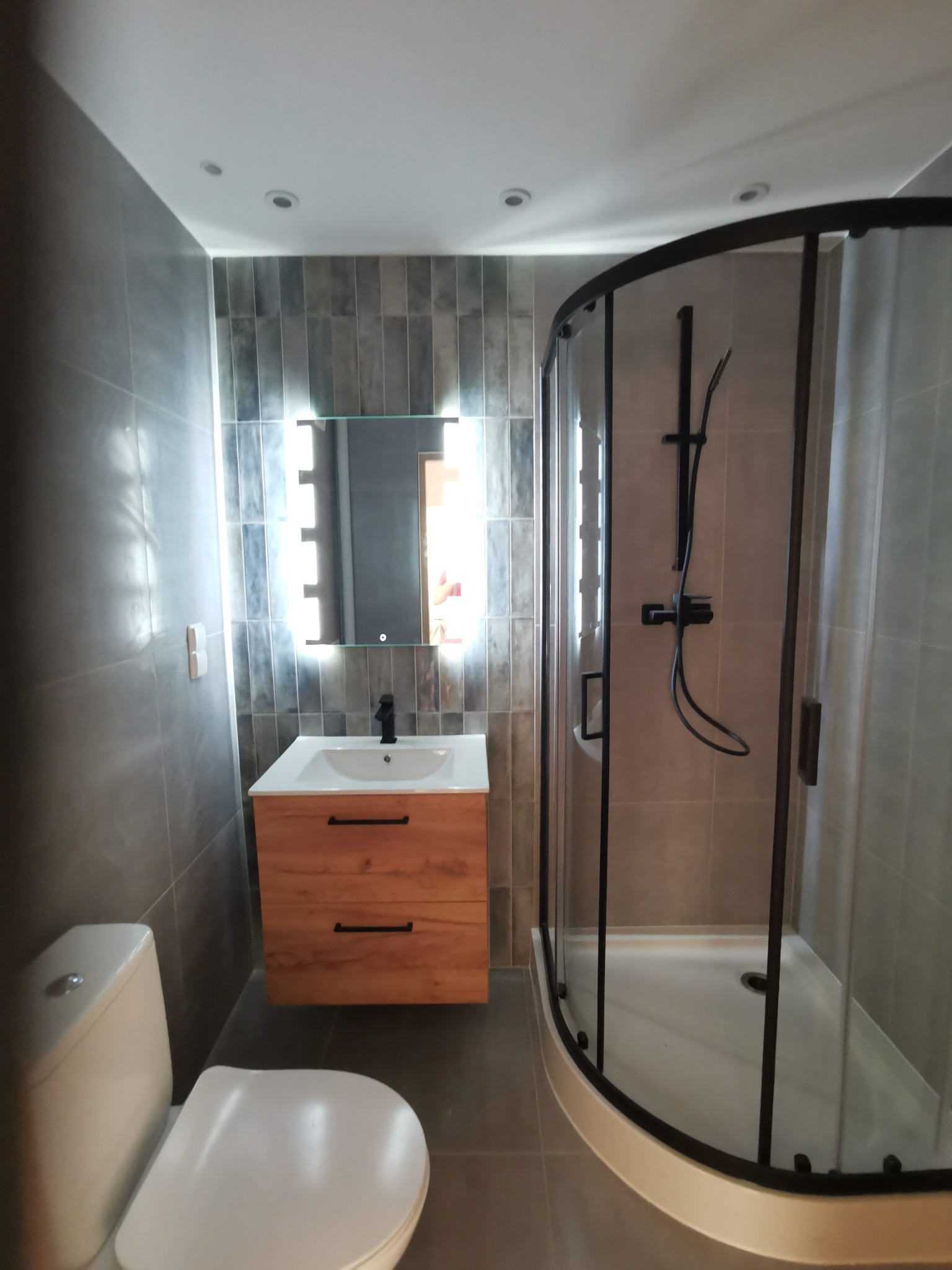 Nowoczesna łazienka z szarymi płytkami, kabiną prysznicową z czarnymi profilami, podświetlanym lustrem nad drewnianą szafką z umywalką i wiszącą toaletą.