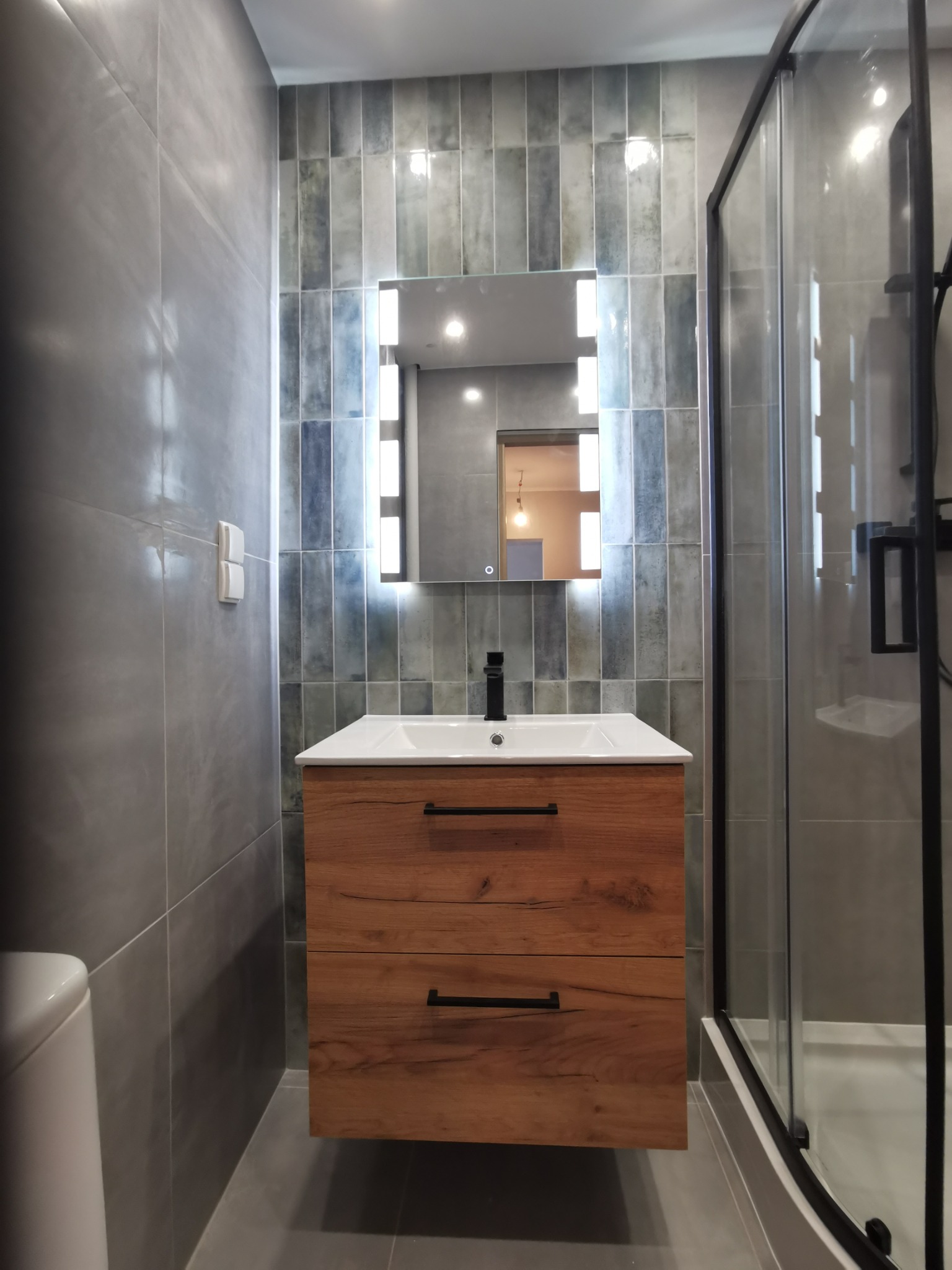 Nowoczesna łazienka w Opolu po renowacji: drewniana szafka pod umywalką z czarną armaturą, podświetlane lustro i kabina prysznicowa z czarnymi profilami na tle szaro-niebieskich płytek.