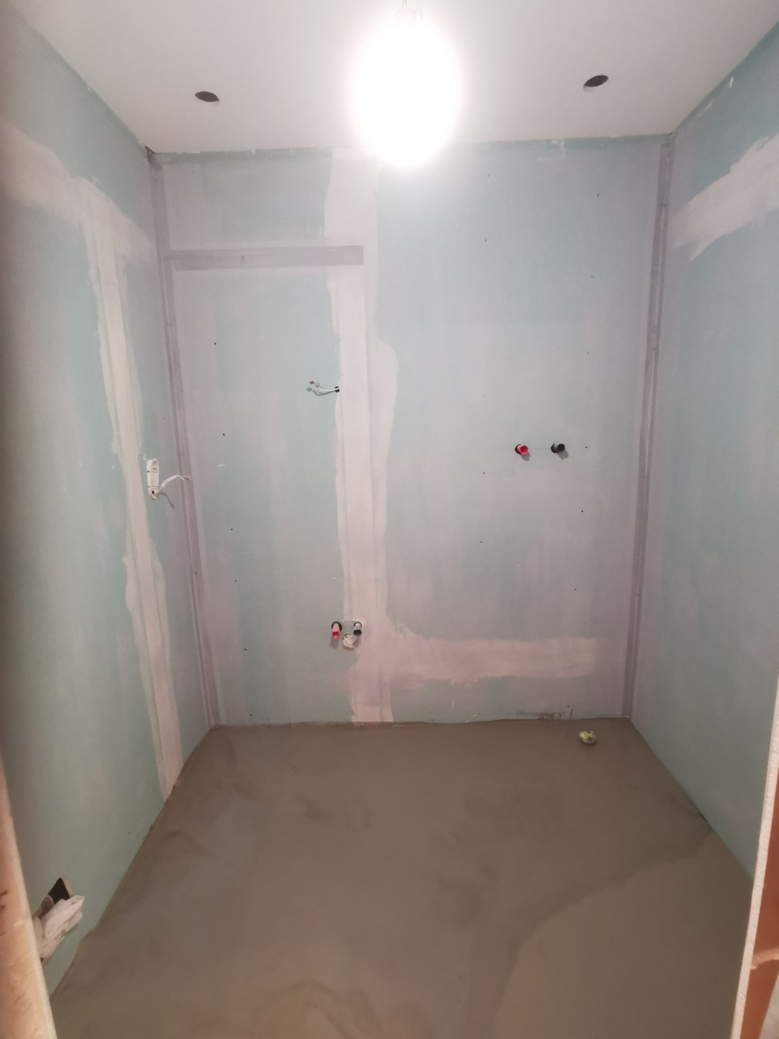 Surowa łazienka w trakcie remontu: ściany z płyt karton-gips, świeżo wylana wylewka podłogowa, widoczne instalacje wodne i elektryczne.