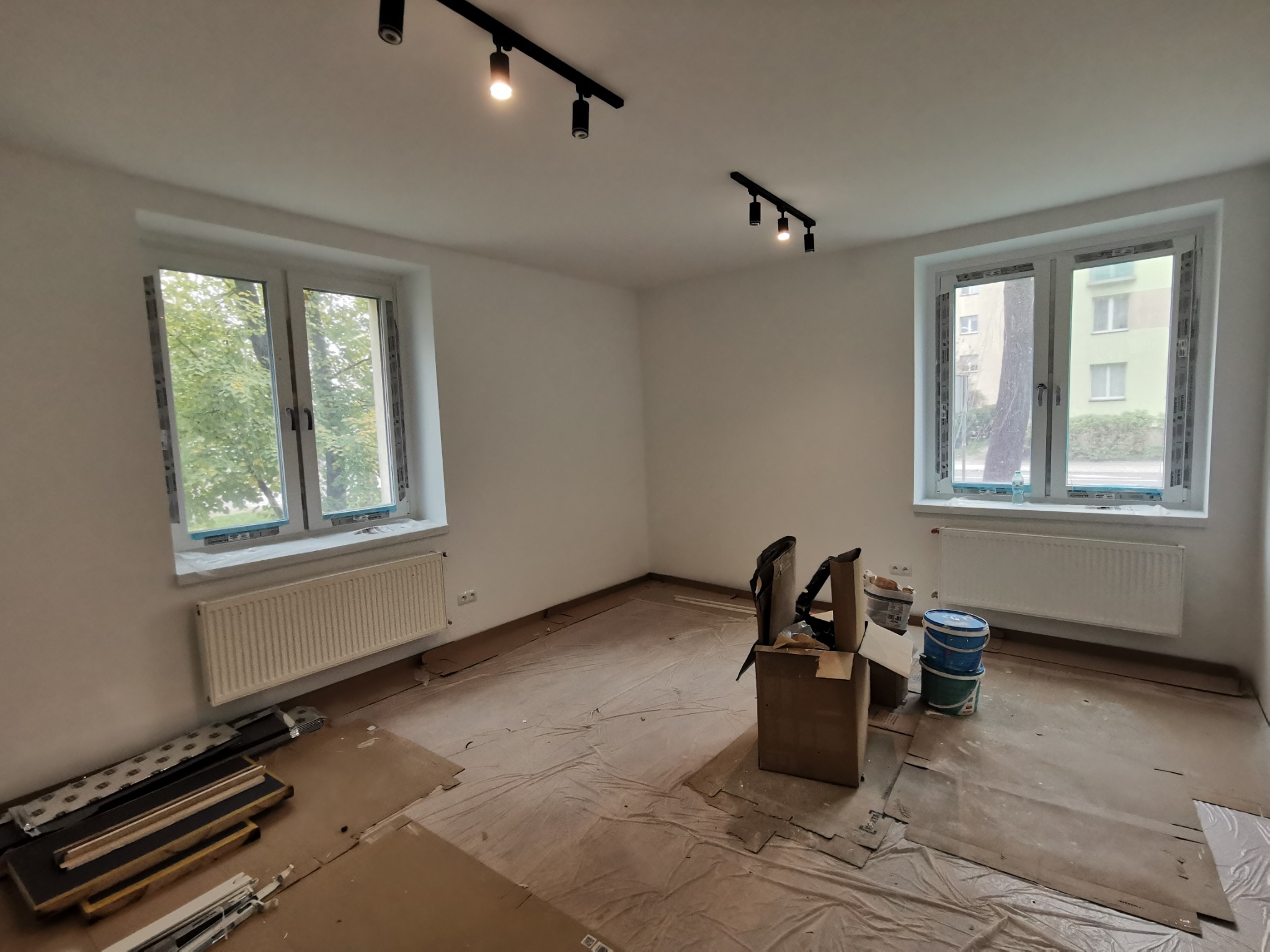 Pokój w trakcie przygotowań do prac wykończeniowych, zabezpieczona podłoga folią malarską, widoczne narzędzia i puste pojemniki po farbach, czarne reflektory na suficie.