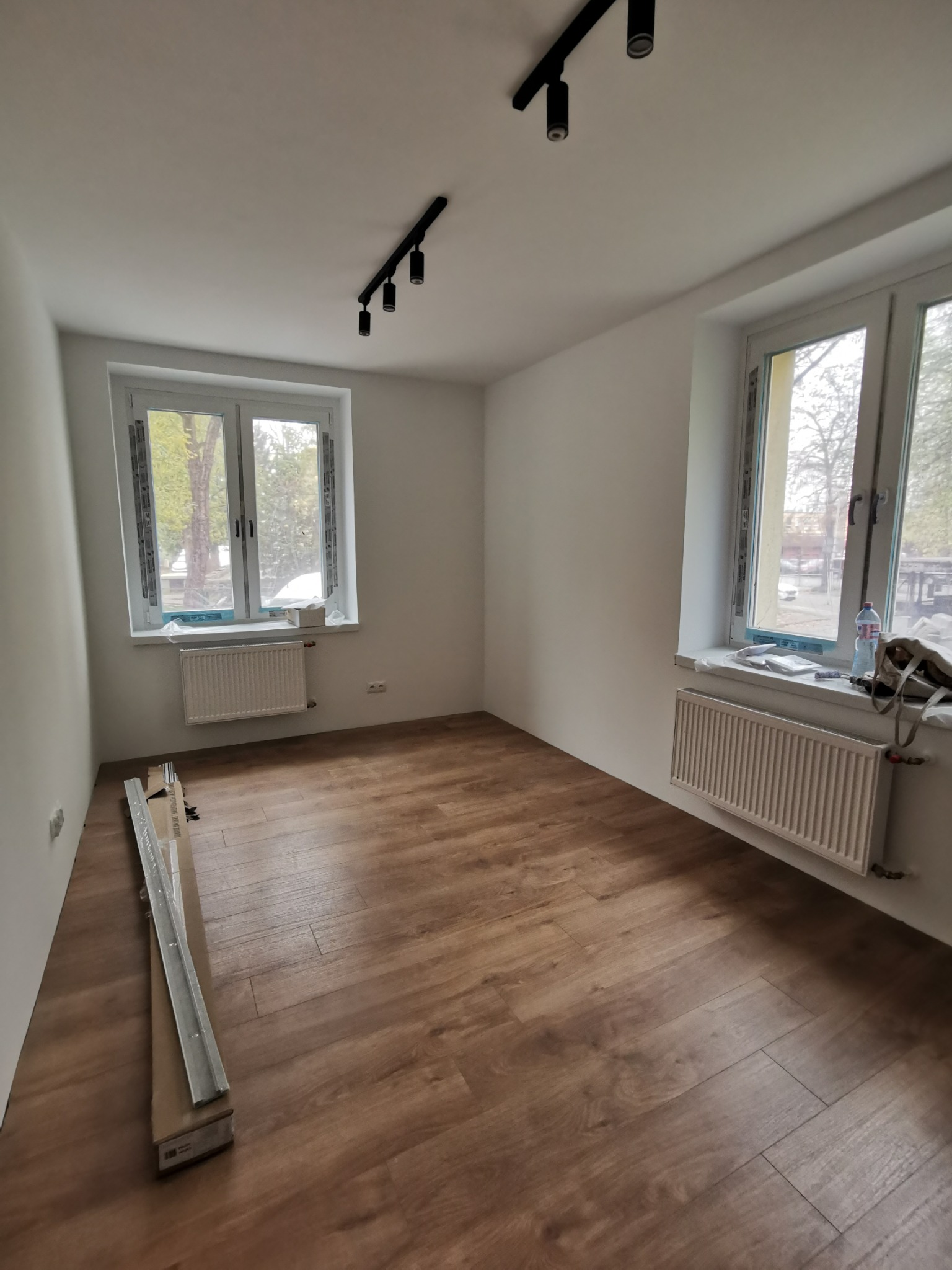 Pusty pokój po remoncie z panelami podłogowymi w odcieniach brązu, dwa okna z białymi ramami, grzejniki pod oknami, czarne reflektory na suficie i listwy przypodłogowe w kartonie.