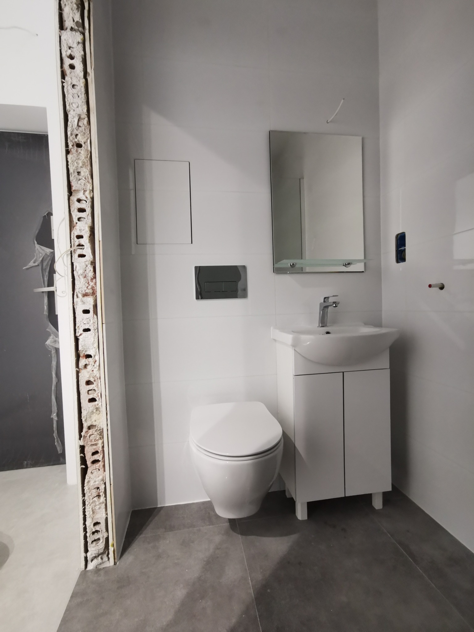 Wnętrze łazienki w trakcie remontu: widoczna toaleta, umywalka z szafką, lustro na tle białych płytek oraz odsłonięta cegła w miejscu niedokończonych prac wykończeniowych.