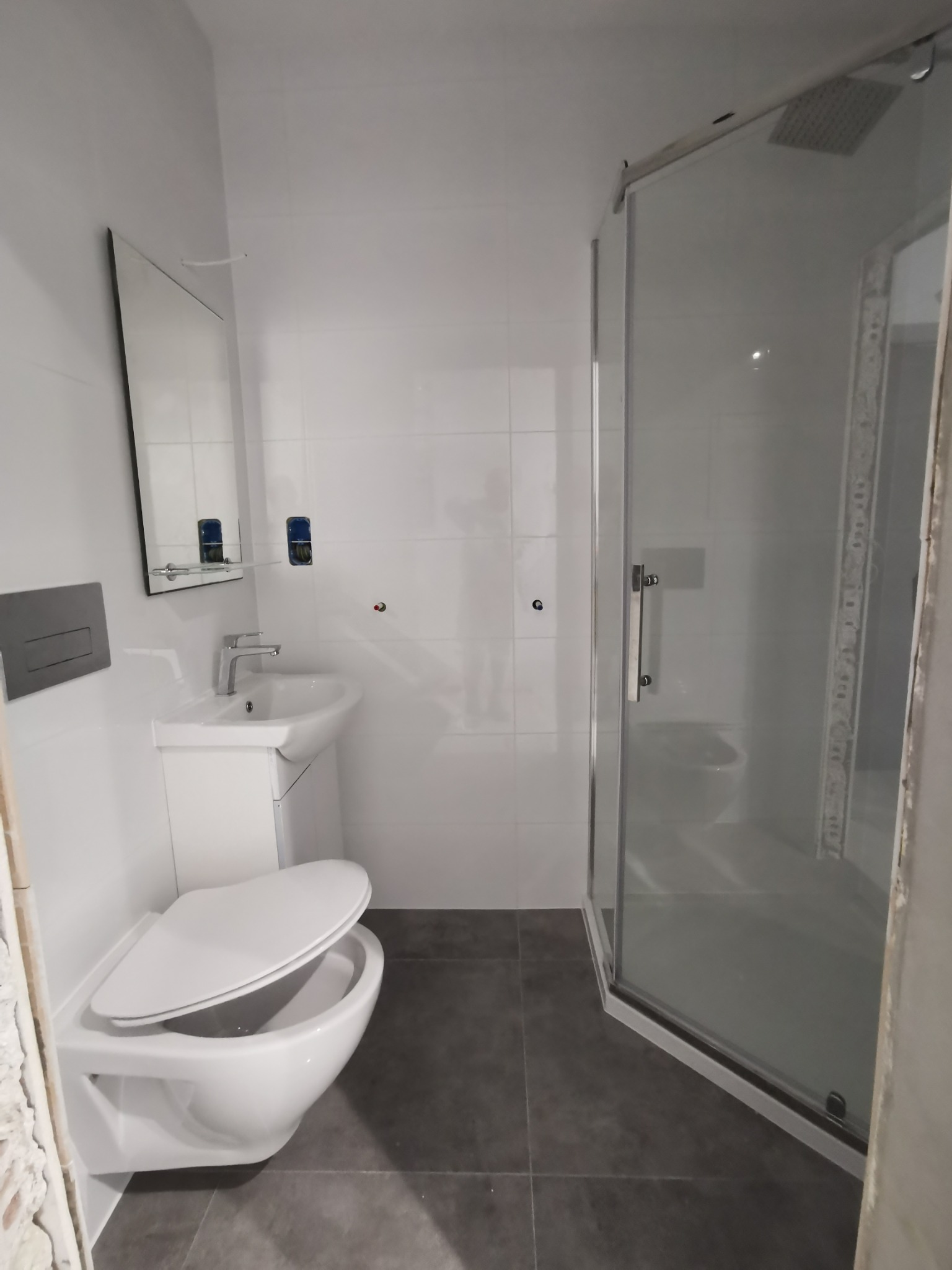 Nowoczesna, jasna łazienka z wiszącą toaletą z otwartą deską, umywalką na szafce, kabiną prysznicową z deszczownicą i szarą podłogą z płytek.