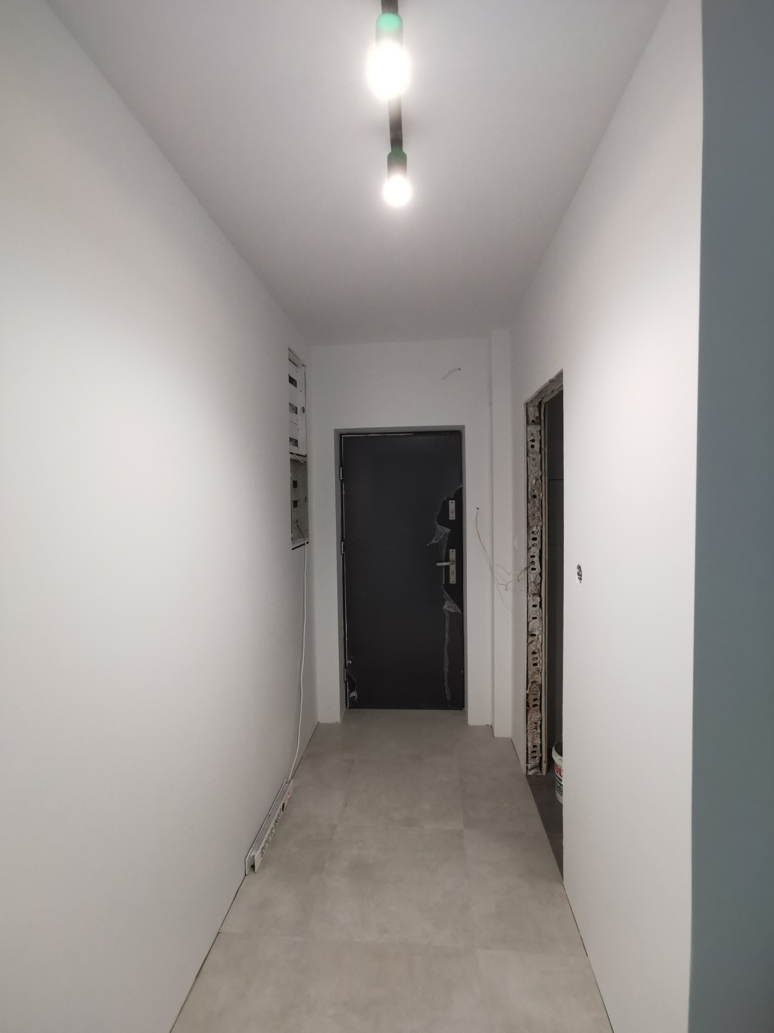 Korytarz w trakcie prac wykończeniowych: świeżo pomalowane białe ściany, szara podłoga, widoczne kable elektryczne i puszka instalacyjna, surowa ościeżnica bez drzwi i czarne drzwi wejściowe...