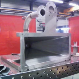 Konstrukcja z pospawanych elementów metalowych o złożonym kształcie, z widocznymi spawami i otworami montażowymi, umieszczona na perforowanym stole warsztatowym w hali produkcyjnej.