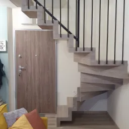 Drewniane schody spiralne z czarną balustradą, widok z dołu, obok drzwi wejściowych i żółtej sofy