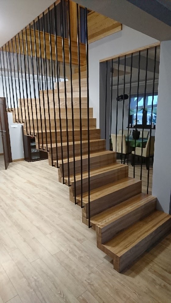 Nowoczesne schody z jasnego drewna z czarnymi, pionowymi balustradami, widoczne z dołu, z elementami wyposażenia wnętrza w tle.