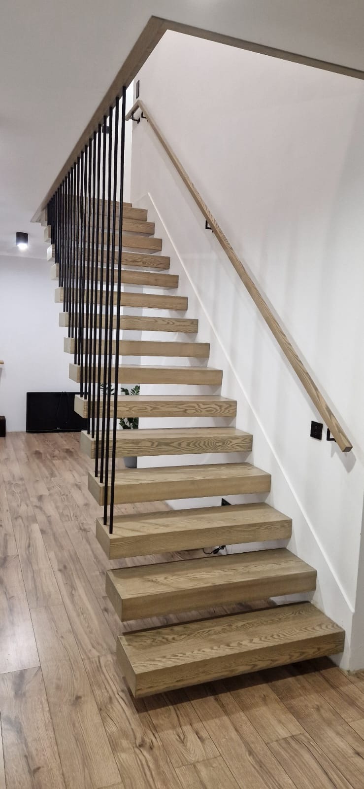 Nowoczesne schody z jasnego drewna z czarnymi, pionowymi balustradami i drewnianą poręczą na białej ścianie, widoczne z dołu w kierunku góry.