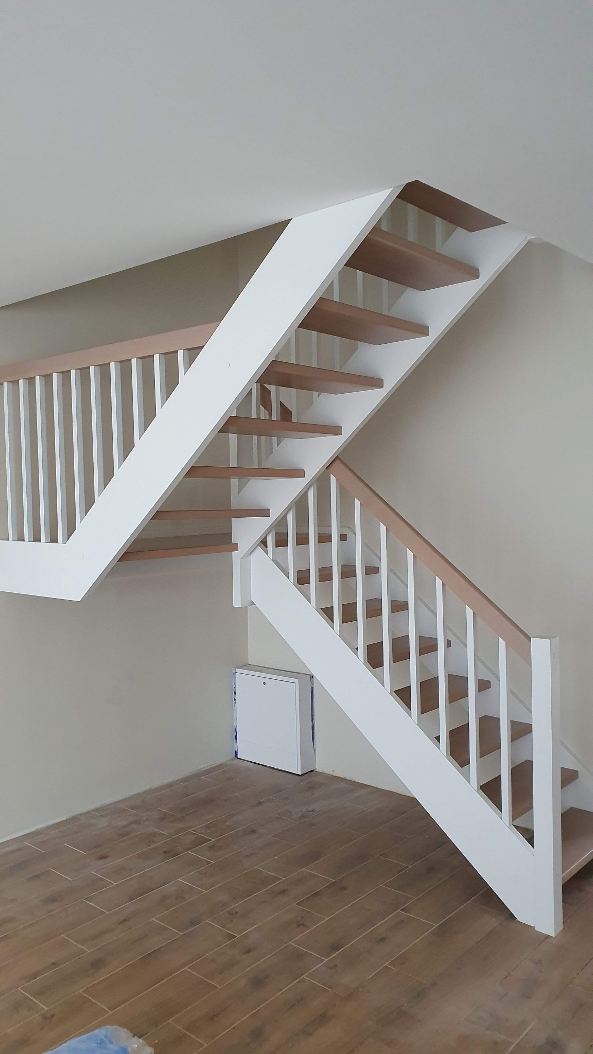 Nowoczesne schody z drewnianymi stopniami i białymi barierkami, widok z dołu na tle beżowej ściany i podłogi imitującej drewno.