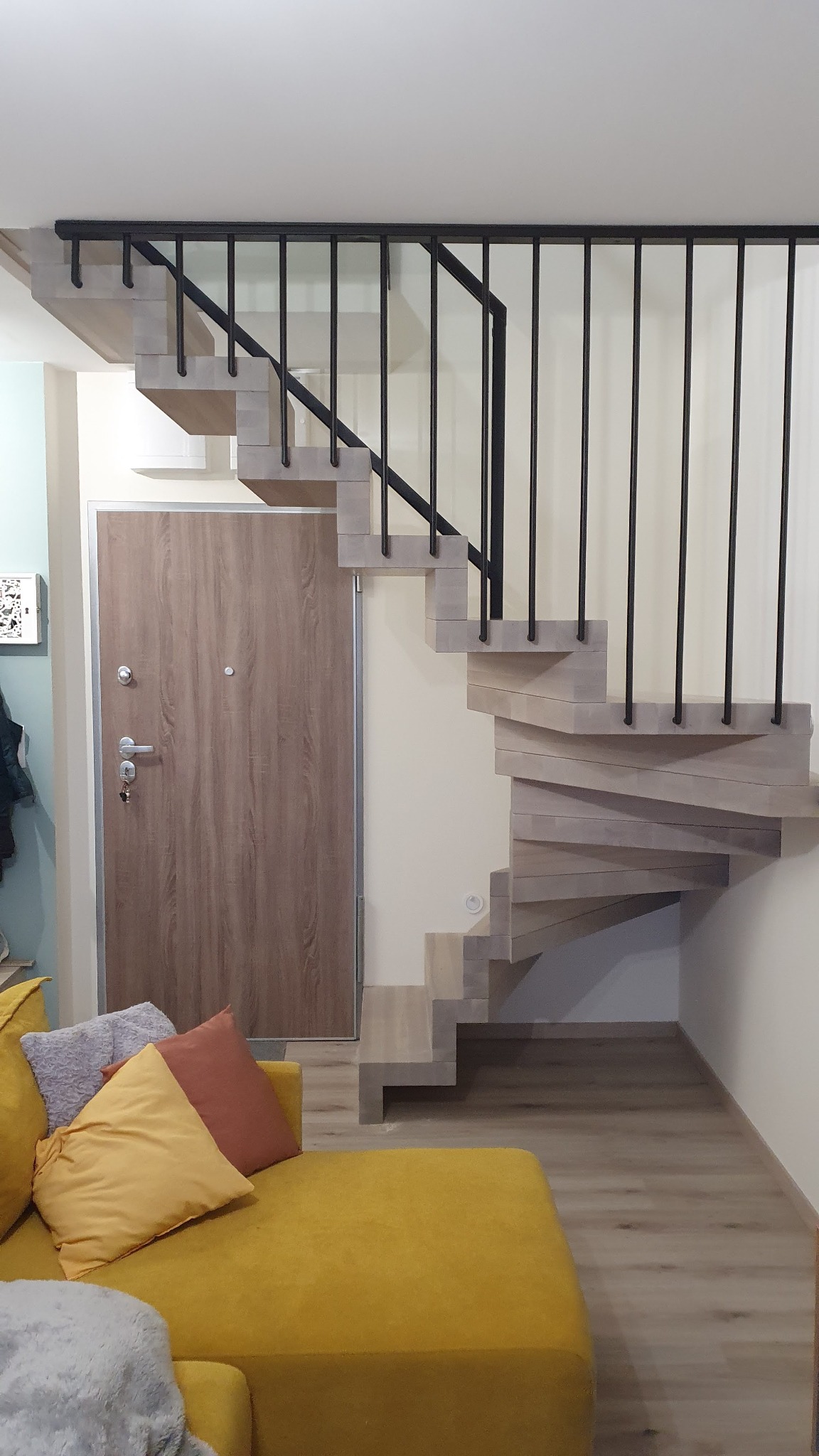 Drewniane schody spiralne z czarną balustradą, widok z dołu, obok drzwi wejściowych i żółtej sofy