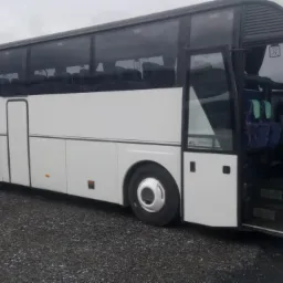 Biały autokar turystyczny z otwartymi drzwiami wejściowymi, widok z boku na szarym żwirowym podłożu i pochmurnym tle.