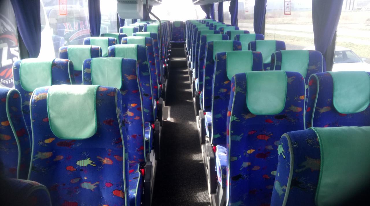 Wnętrze autobusu z rzędami niebieskich foteli z kolorowym wzorem i jasnymi zagłówkami, widok wzdłuż korytarza.