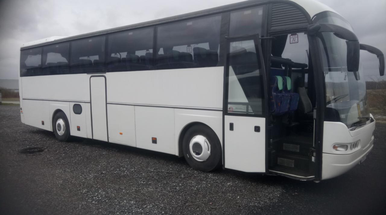 Biały autokar turystyczny z otwartymi drzwiami wejściowymi, widok z boku na szarym żwirowym podłożu i pochmurnym tle.