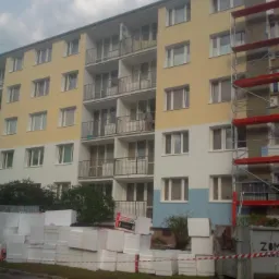 Blok mieszkalny w trakcie prac termomodernizacyjnych, widoczne rusztowania, ocieplenie ze styropianu na ziemi, osoba na balkonie.