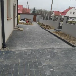 Ułożona, szara kostka brukowa na podjeździe przed domem, widoczne obrzeża, w tle budynek gospodarczy i ogrodzenie.