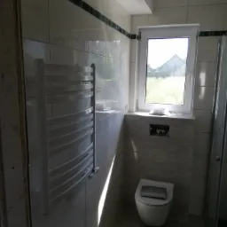 Nowoczesna łazienka z białymi płytkami, grzejnikiem drabinkowym, podwieszaną toaletą z chromowanym przyciskiem oraz oknem z widokiem na zieleń.