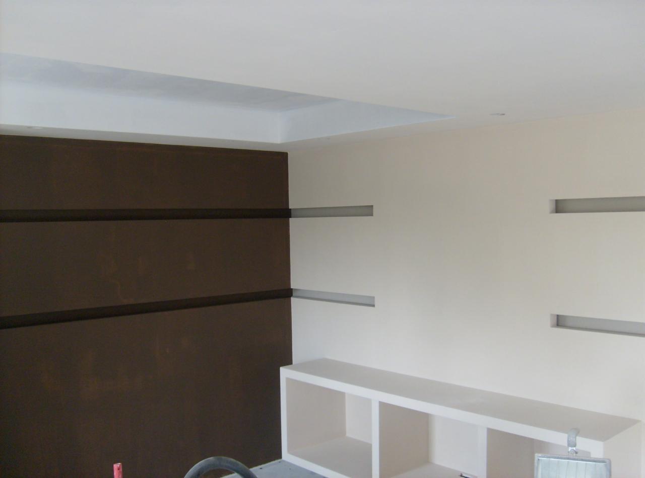 Wnętrze pokoju w trakcie malowania: brązowa ściana z poziomymi paskami, kremowa ściana z wnękami, biały regał pod ścianą. Widoczne narzędzia malarskie.