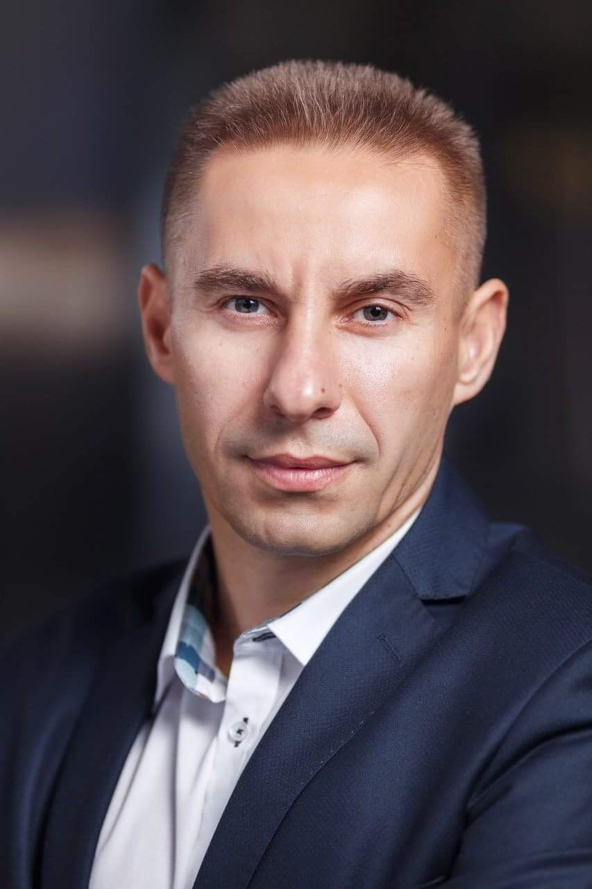 Portret mężczyzny w ciemnym garniturze i białej koszuli z niebieskimi akcentami, patrzącego wprost, na ciemnym tle.