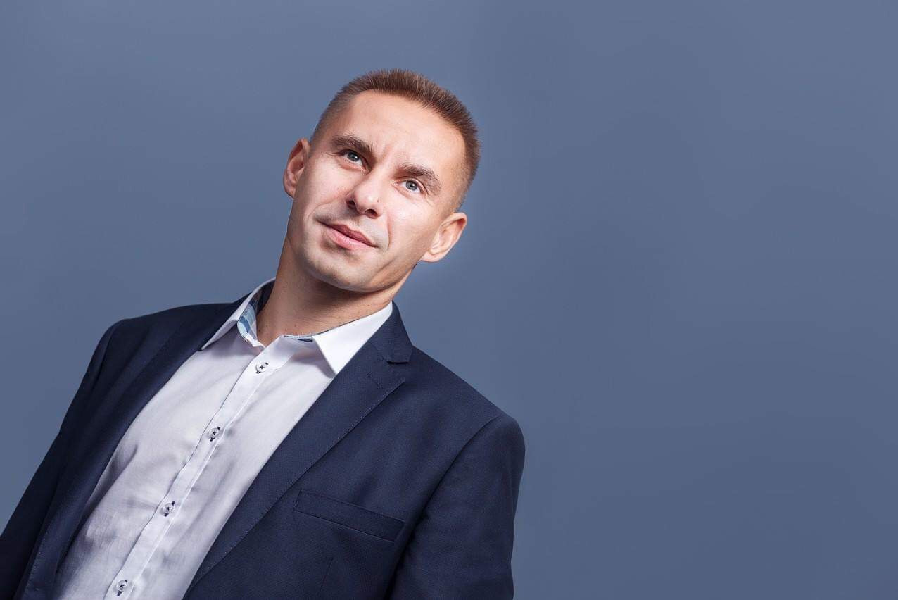 Portret mężczyzny w ciemnym garniturze i białej koszuli, spoglądającego w górę w lewo na szarym tle.