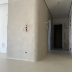 Wnętrze w trakcie prac wykończeniowych: ściany pokryte szarą, nakrapianą fakturą, widoczne wystające kable elektryczne i puszki instalacyjne, podłoga również w szarym odcieniu, surowe drzwi w tle.