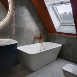 Nowoczesna łazienka na poddaszu z białą wanną wolnostojącą, umywalką na szafce i toaletą. Uwagę przykuwają zielono-miedziane płytki i okno dachowe.