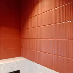 Fragment łazienki z terakotowymi płytkami ściennymi o pionowych żłobieniach i gładkimi płytkami w rogu, dolna część ściany wykończona płytkami imitującymi lastryko, widoczny czarny panel spłuczki.