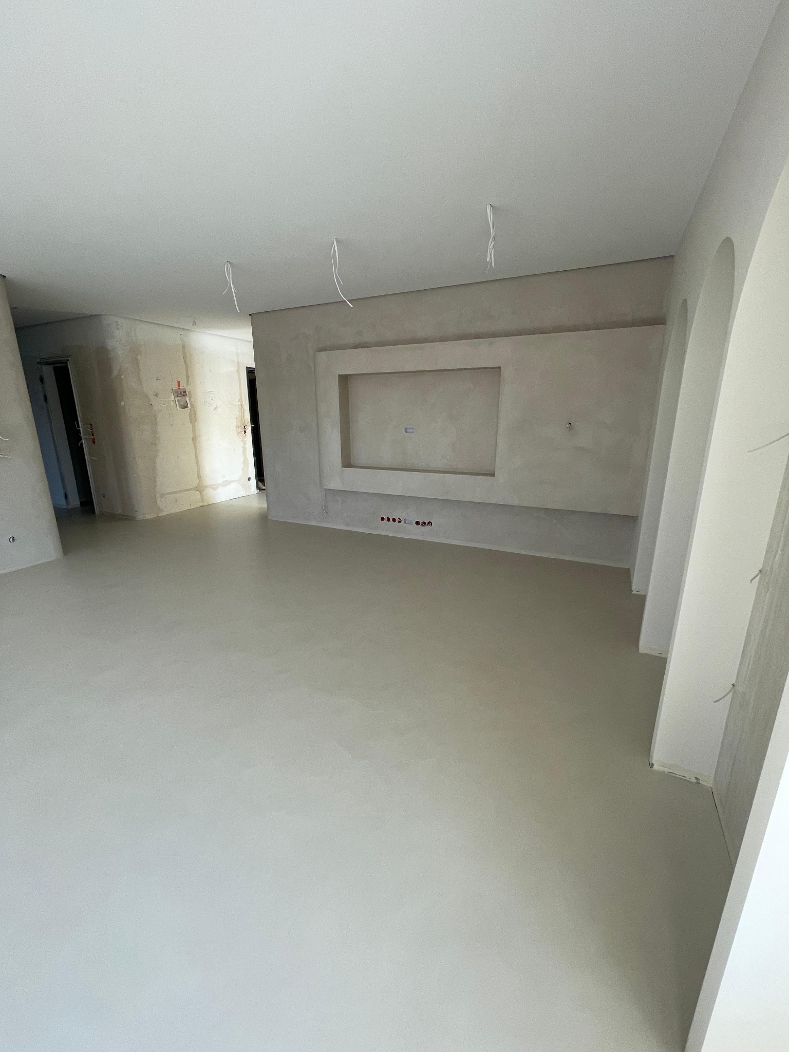 Wnętrze w trakcie prac wykończeniowych: gładka, jasna podłoga, ściany z elementami dekoracyjnymi i instalacją elektryczną, surowe wykończenie w sąsiednim pomieszczeniu, wiszące kable.
