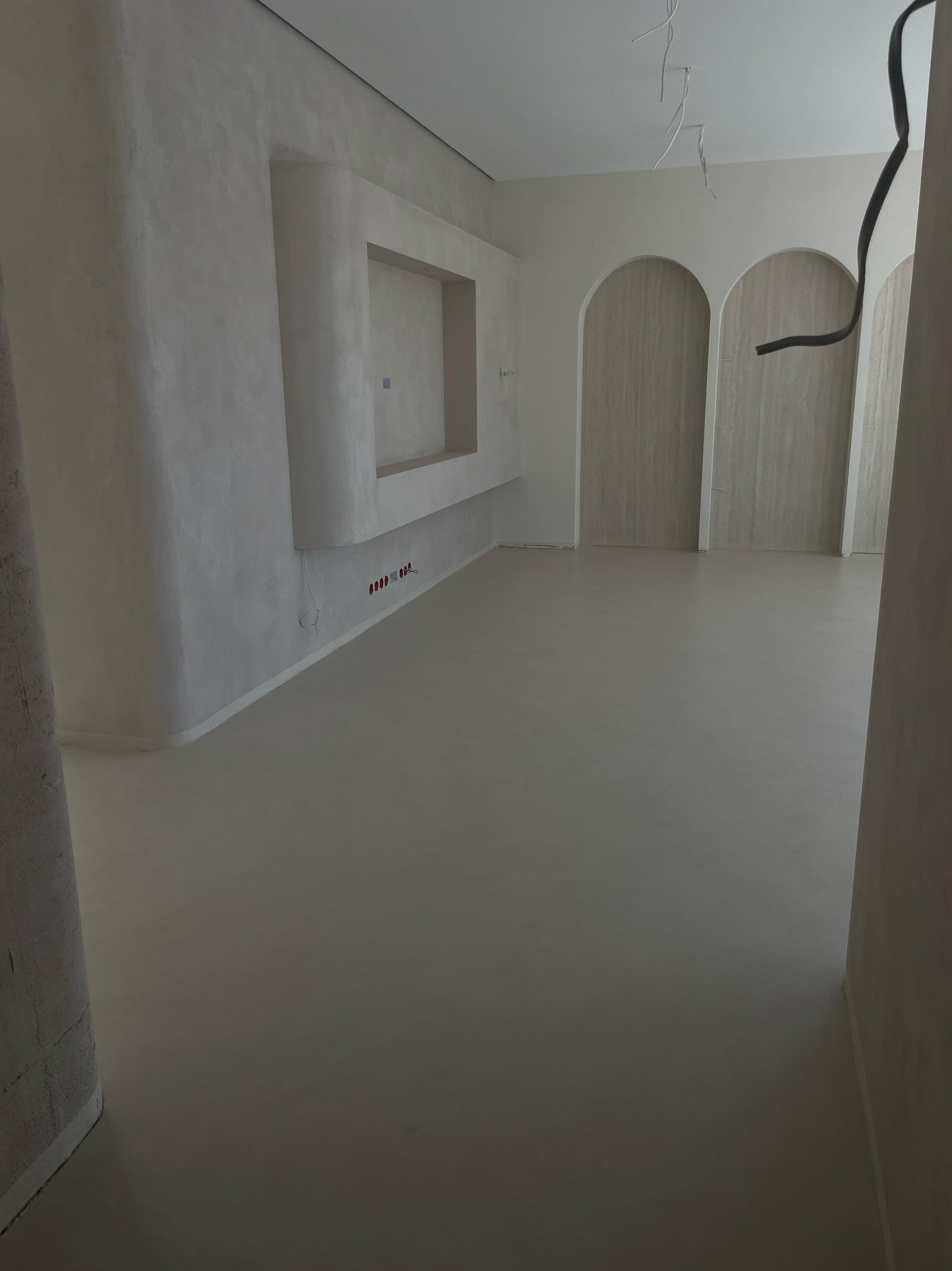 Wnętrze w trakcie prac wykończeniowych: gładkie, jasne ściany z wnęką, podłoga w jednolitym kolorze, widoczne kable i otwory na gniazdka, drzwi z łukowym nadprożem.