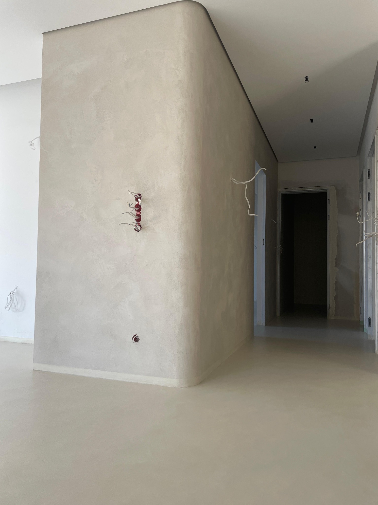 Wnętrze w trakcie prac wykończeniowych: ściany pokryte szarą, nakrapianą fakturą, widoczne wystające kable elektryczne i puszki instalacyjne, podłoga również w szarym odcieniu, surowe drzwi w tle.