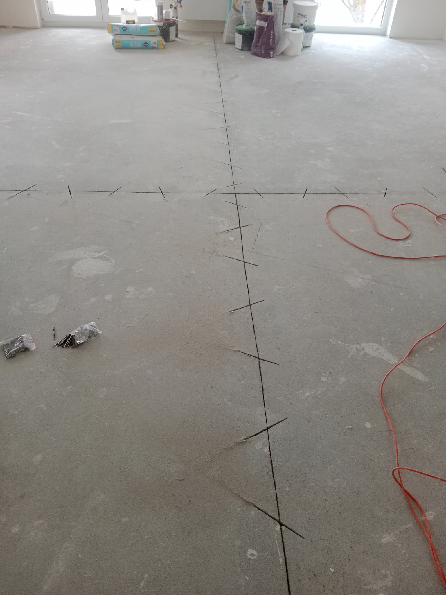Przygotowanie podłoża pod posadzkę: wyznaczone linie cięcia na szarej wylewce, widoczne narzędzia i materiały budowlane w tle.