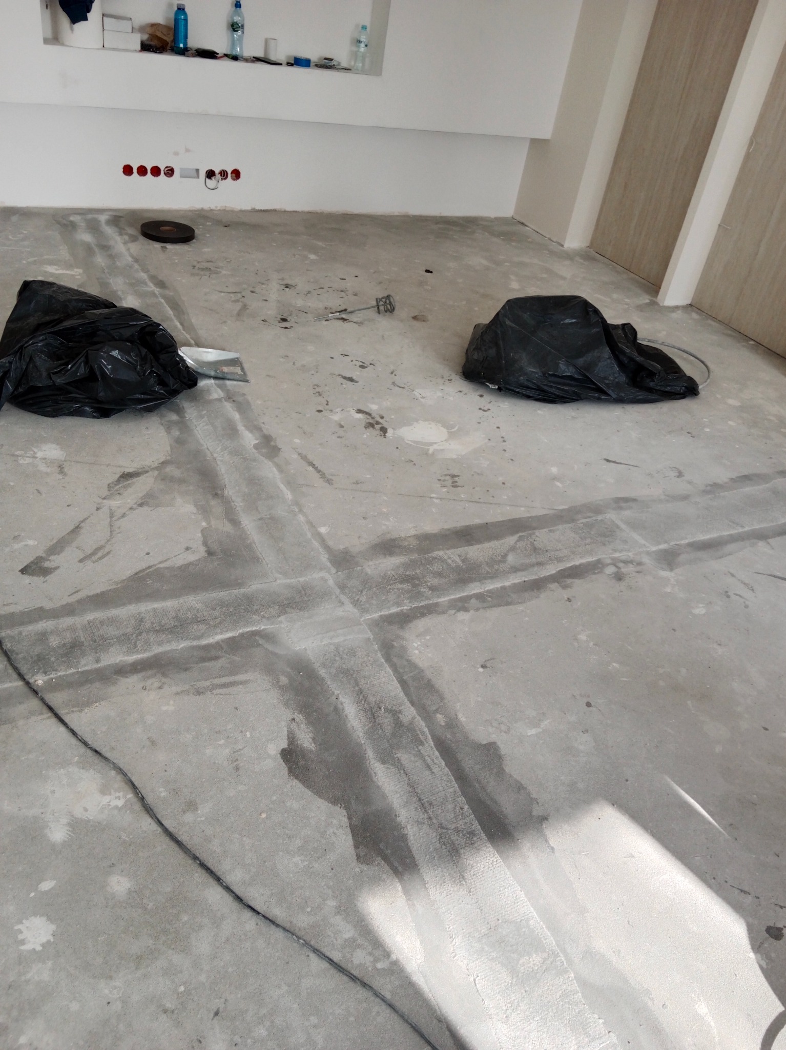 Przygotowanie podłoża pod posadzkę: wyrównywanie wylewki, widoczne ślady po szlifowaniu i wypełnianiu spoin, dwa czarne worki na gruz, przewód elektryczny na podłodze, otwory na gniazdka w ścianie.