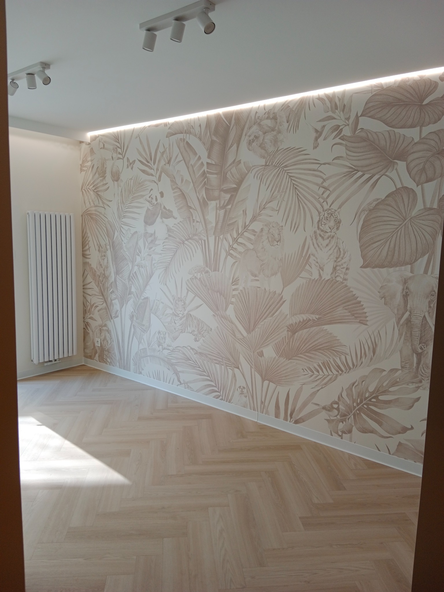 Jasne wnętrze z parkietem w jodełkę i tapetą w motywy dżungli z ukrytymi zwierzętami, oświetlenie LED podsufitowe, biały grzejnik.