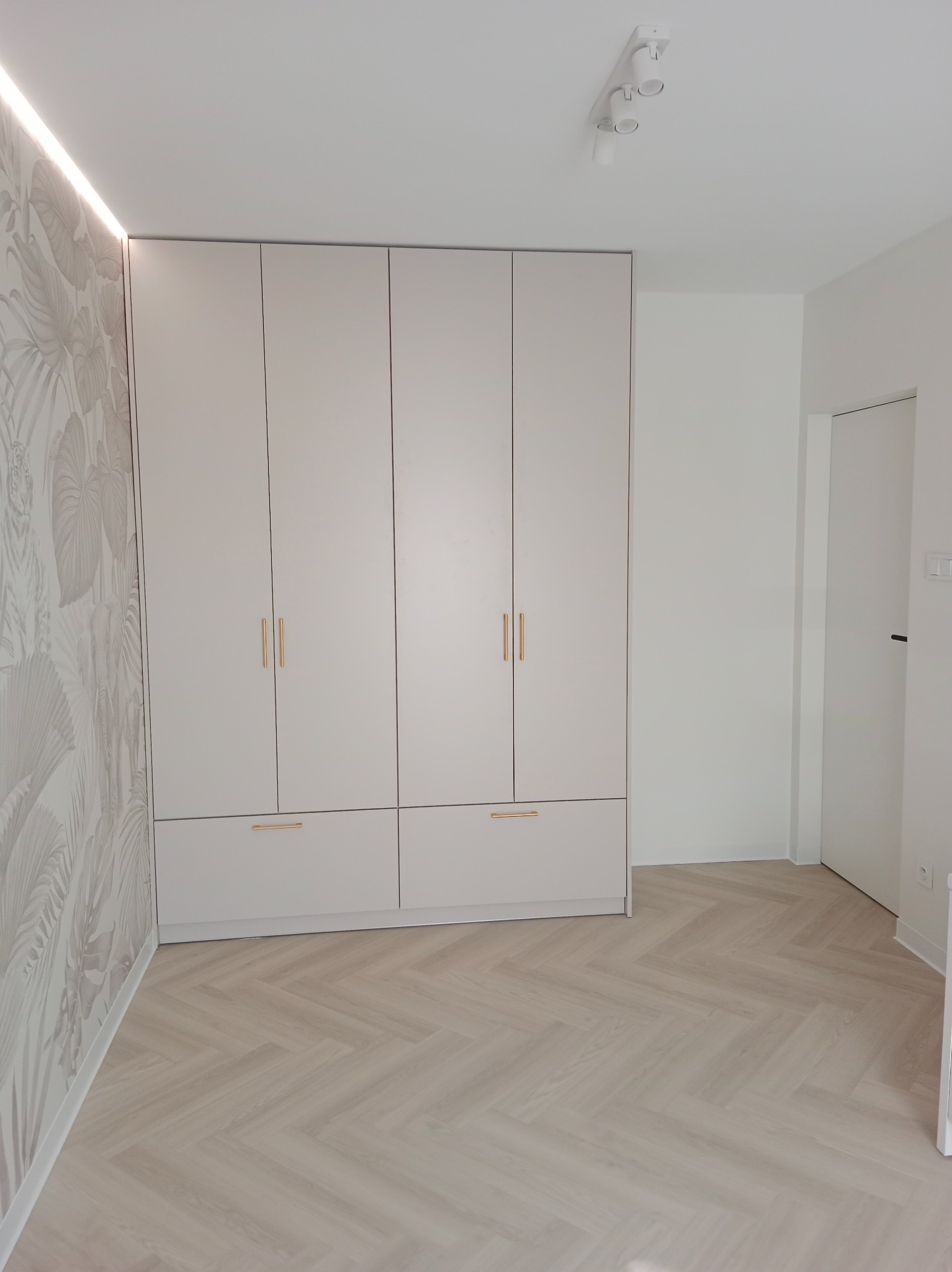 Jasny pokój z czterodrzwiową szafą w zabudowie, parkietem ułożonym w jodełkę i tapetą w tropikalne liście z delikatnym oświetleniem LED.