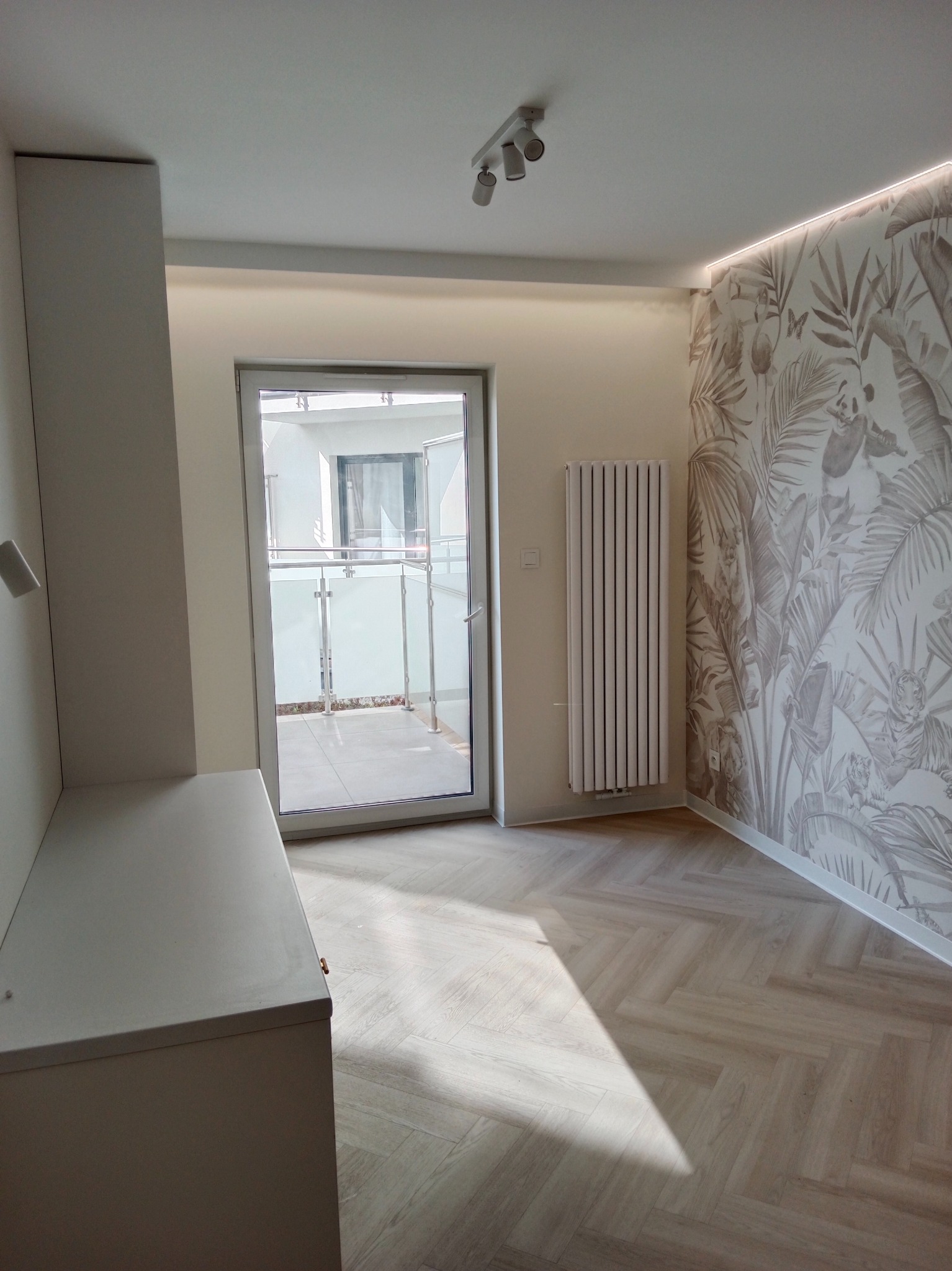 Jasne wnętrze z parkietem w jodełkę, ścianą z tapetą w motywy roślinne, grzejnikiem panelowym i wyjściem na balkon.