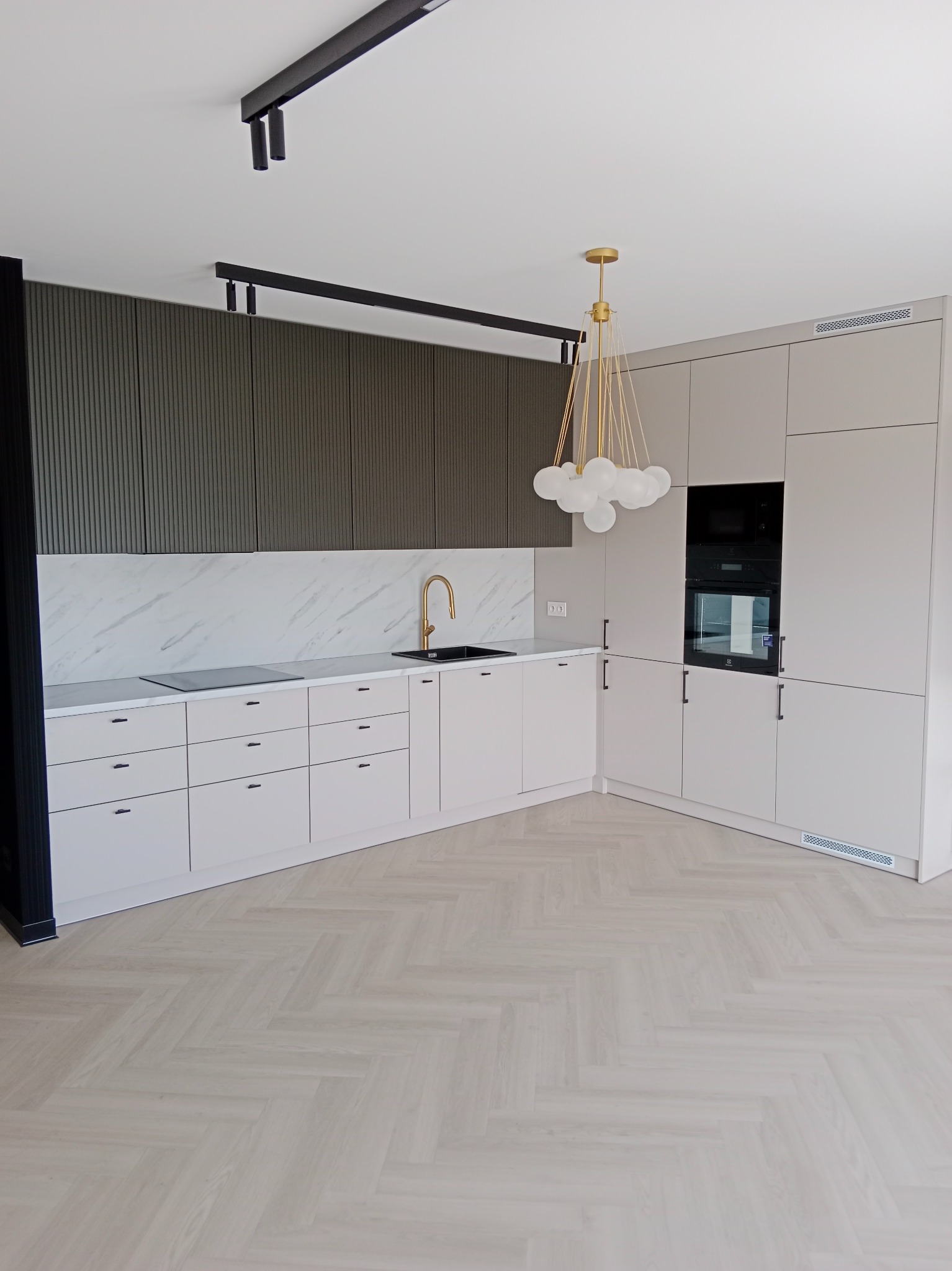 Nowoczesna kuchnia z beżowymi szafkami, złotą armaturą i marmurowym blatem, oświetlona lampą z białymi kloszami i czarnymi szynami sufitowymi, na podłodze parkiet ułożony w jodełkę.
