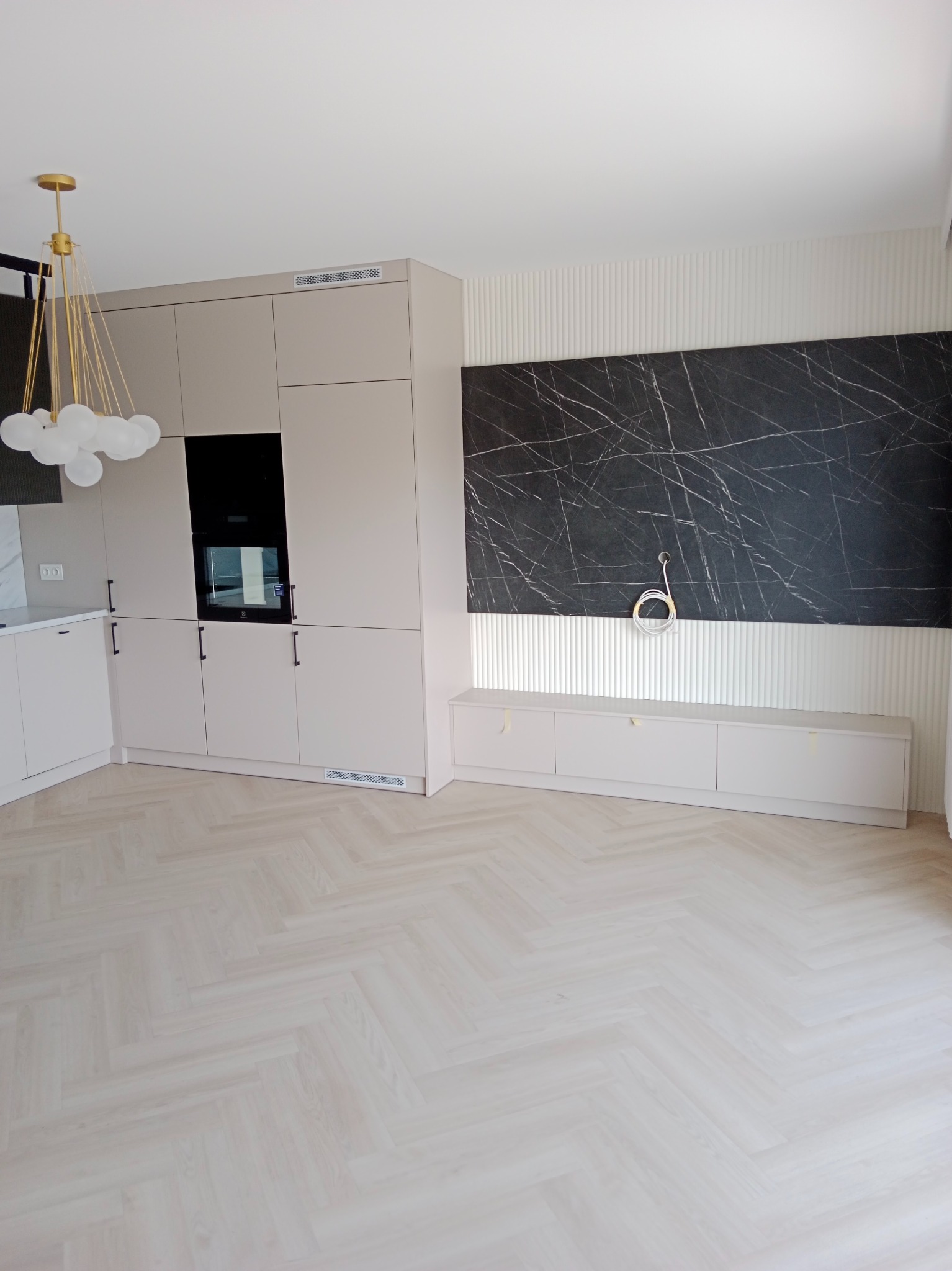 Jasne, nowoczesne wnętrze z parkietem w jodełkę, zabudową kuchenną w odcieniach beżu z czarnym piekarnikiem, oraz ścianą z czarnym marmurem i wystającymi kablami.
